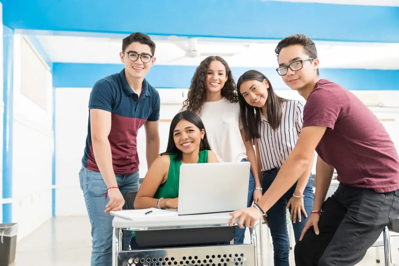 Grupo de estudiantes sonriendo alrededor de una laptop en un salón de clases, representando el aprendizaje colaborativo del Modelo Dual